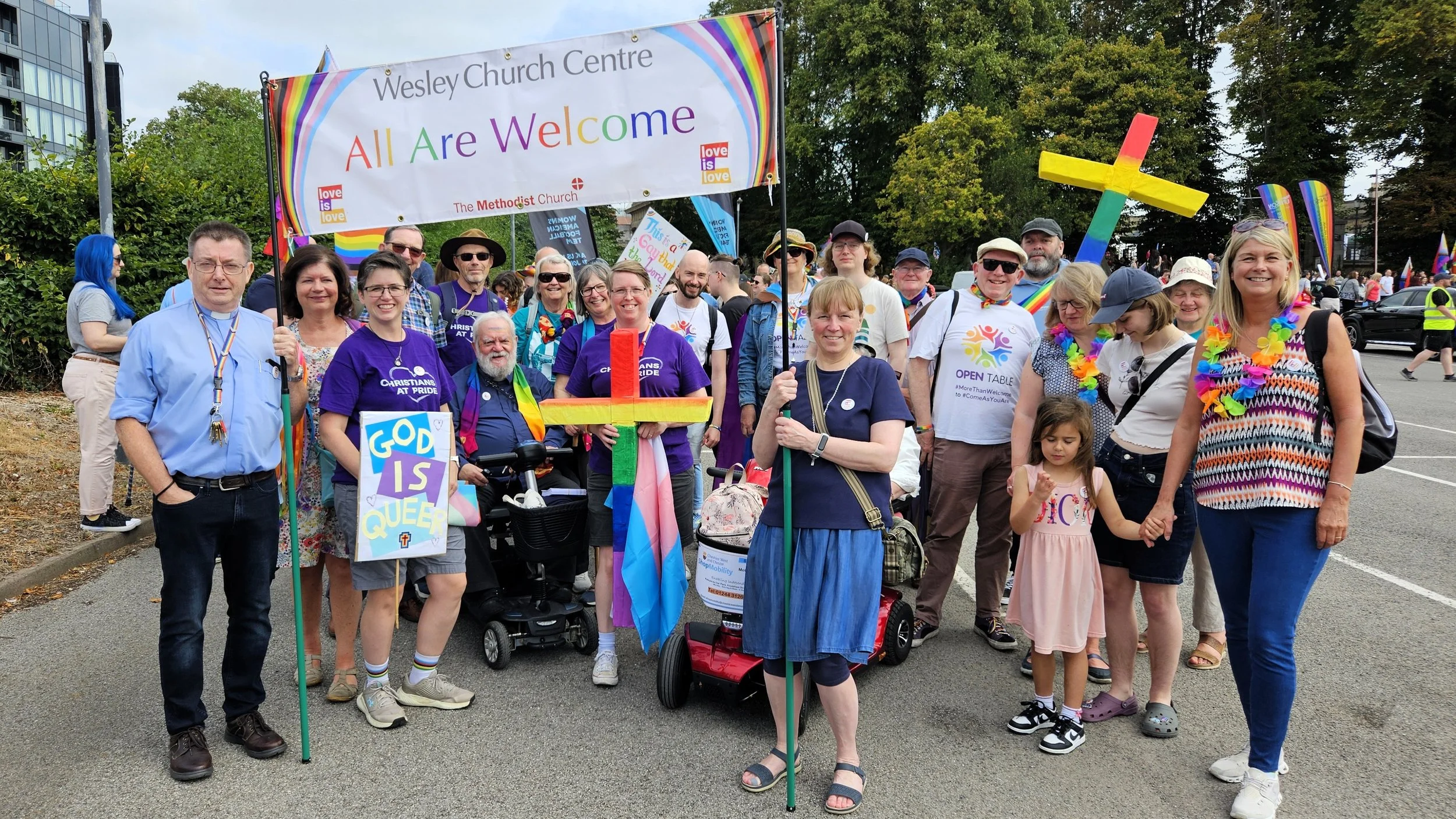 Members of the Wesley Church Centre and Open Table Chester community preparing to march in the city’s Pride parade with a banner saying ‘All Are Welcome’. Two are carrying crosses painted in rainbow colours.